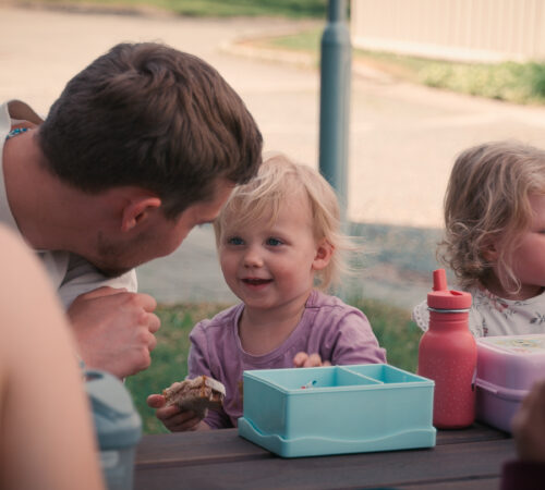 Flere barn og voksne sitter å spiser på en benk. En voksen lener seg ned til et av barna. Barnet smiler med en brødskive i hånda.