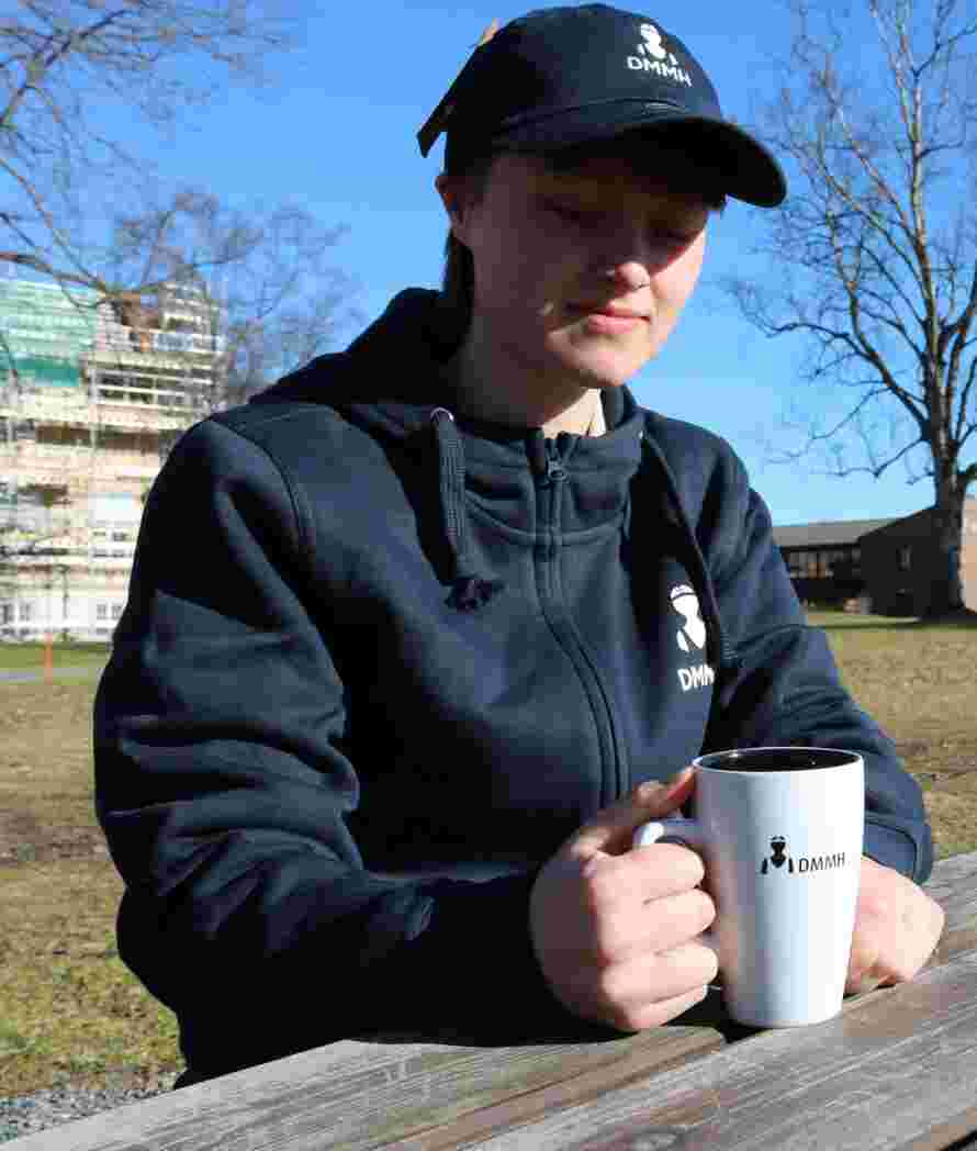Bilde av en hvit og sort DMMH kopp som blir holdt av en student på et bord.